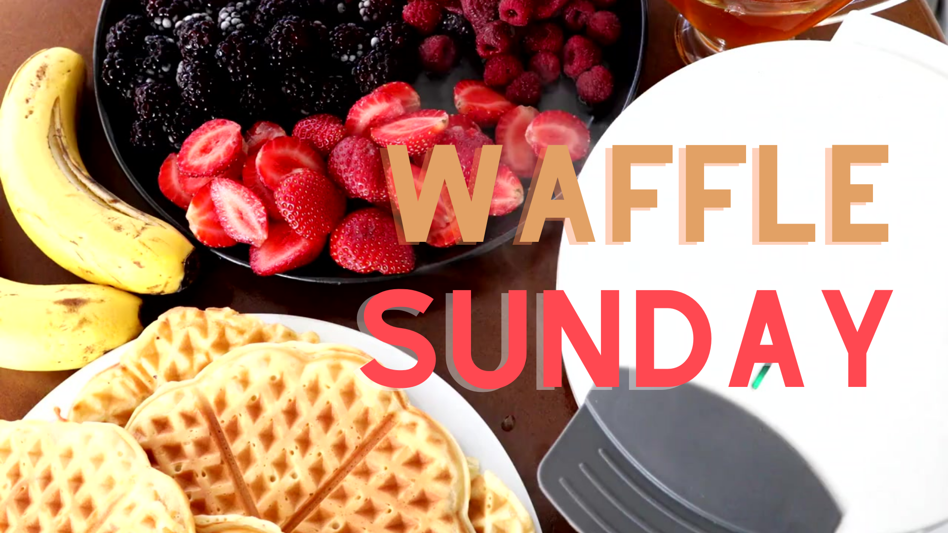 waffle iron surrounded by plate of waffles, bananas, and a plate of strawberries and other berries